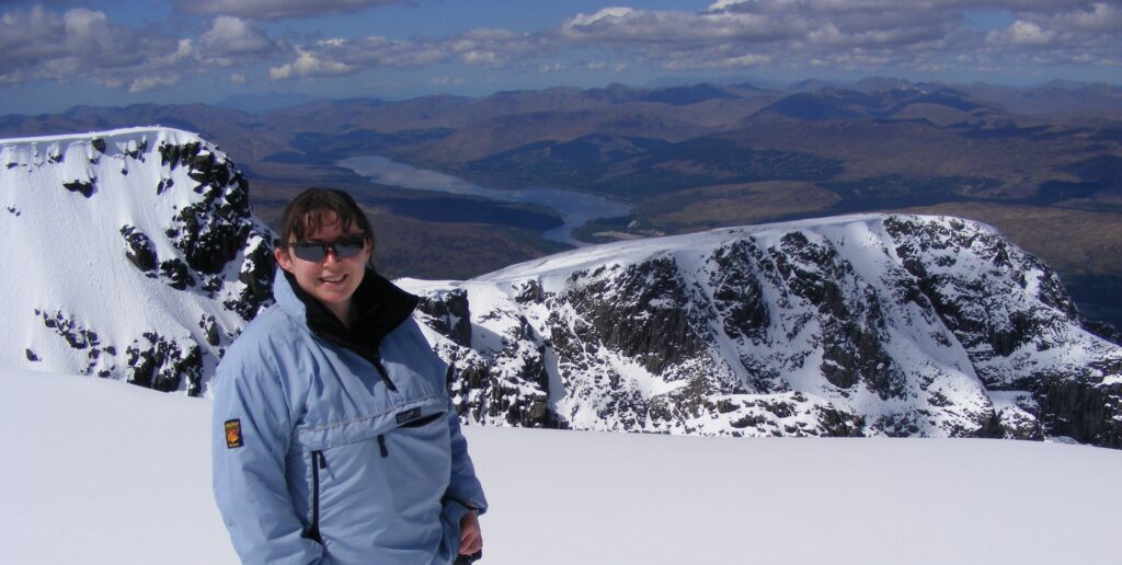 Woman stood on top of Ben Nevis in Scotland. The sun is shining and there is snow on the ground. 