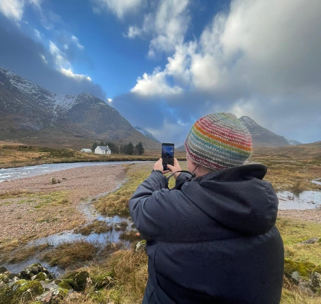 Woman wearing outdoor clothing taking a photo with her phone of Scottish landscape